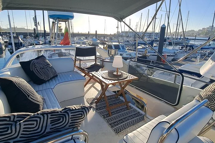 Yacht deck seating with cushions, wooden table, and lamp, moored in a marina.