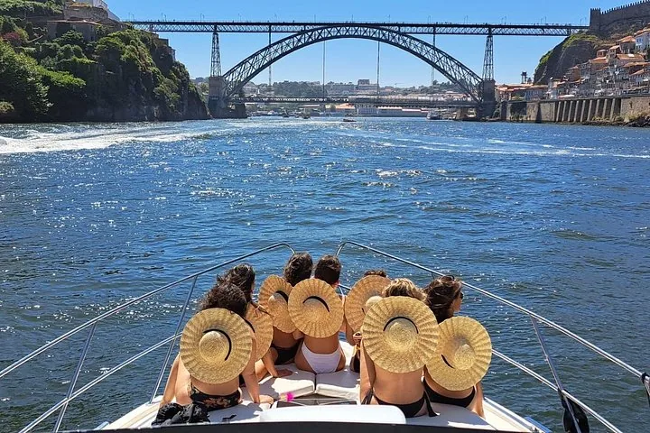 Group of women in straw hats relaxing on yacht bow facing Dom Luís I Bridge.